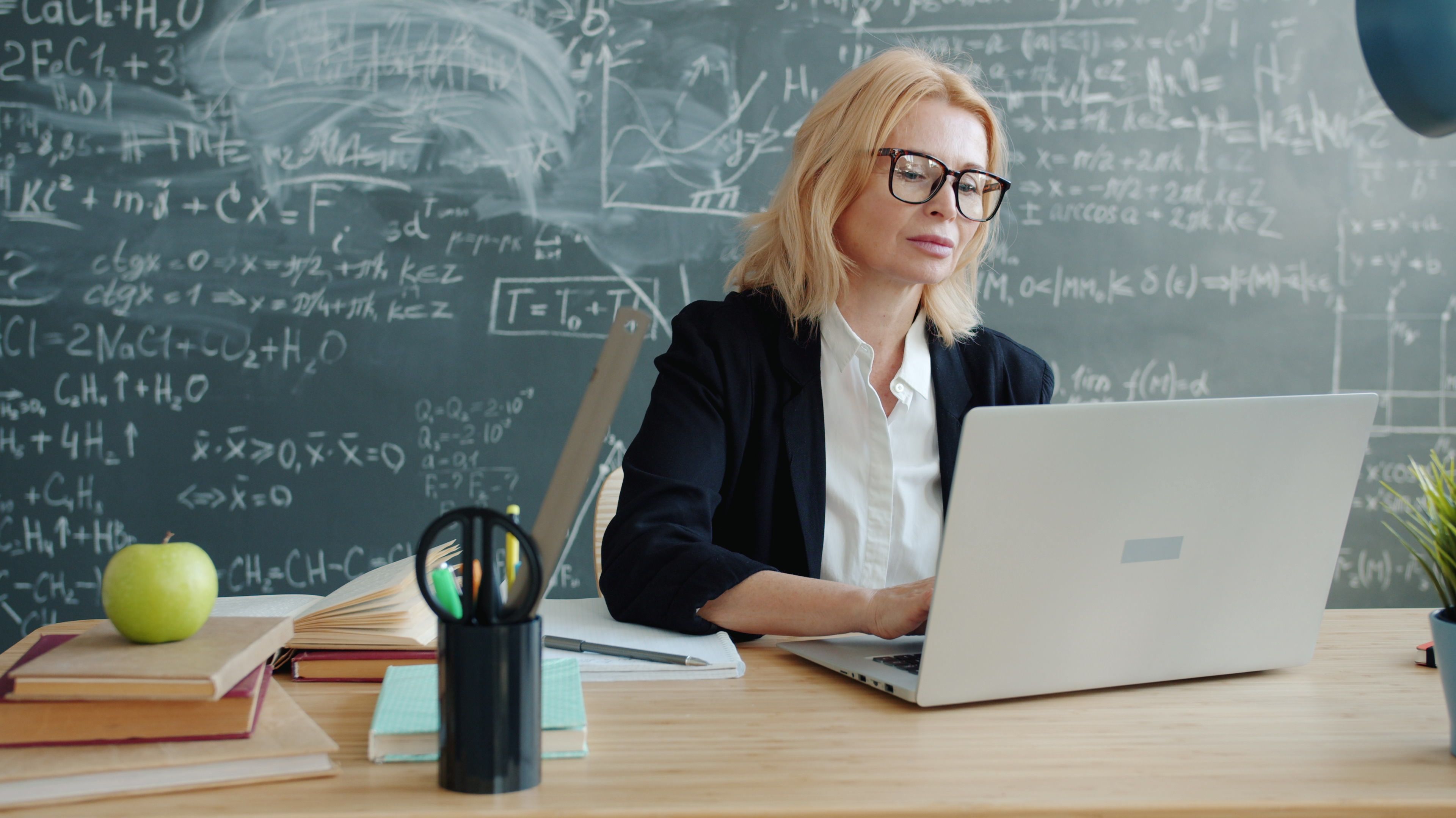 Teacher using a laptop in front of a classroom chalkboard